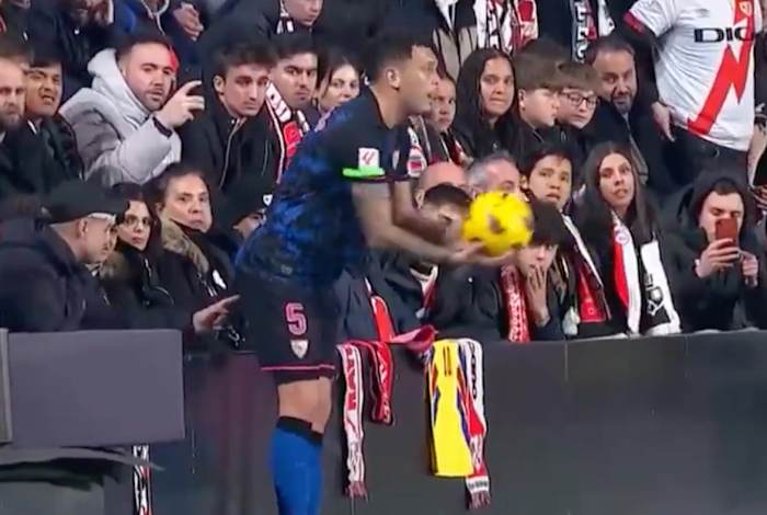 Sevilla's Lucas Ocampos pictured preparing to take a throw-in during a La Liga game against Rayo Vallecano as a fan wearing a baseball cap in the front row appears to poke the player's butt with his index finger